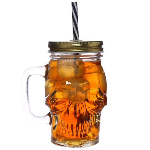 Front view of a skull-shaped glass drinking jar with a golden lid, black and white striped straw, and amber liquid with ice cubes, on a white background.
