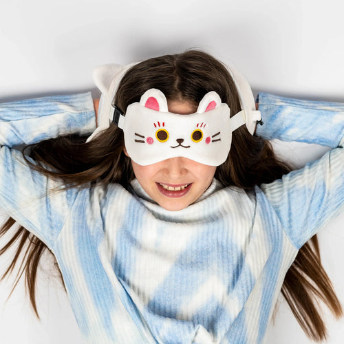 In-context lifestyle shot of a person lying down, wearing a white Maneki Neko lucky cat eye mask with pink ears and embroidered features. A white travel pillow is visible behind their head, and they are wearing a blue and white tie-dye top.