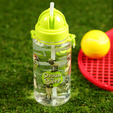 Angled view of a clear children's water bottle with a lime green pop-up straw lid and carrying strap, featuring Shaun the Sheep characters and logo. It sits on green grass, with a red toy racket and yellow ball in the background.