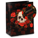 Front-angled product view of a black gift bag featuring a central skull graphic surrounded by vibrant red roses and green vines. The bag is patterned with subtle red roses and has black rope handles with a decorative circular 'Skulls and Roses' tag attached.