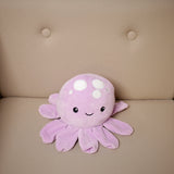 Lilac plush octopus cushion with a smiling embroidered face, white circular spots, and multiple arms, positioned on a beige sofa.