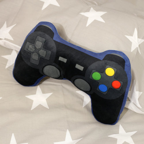 Angled view of a black plush game controller cushion with blue trim, showcasing colourful green, yellow, blue, and red buttons, a grey directional pad, and grey analogue stick details, resting on a grey fabric with white stars.