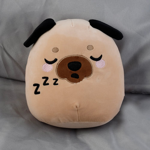 Angled view of a sleeping light brown pug plush toy with pink cheeks, a brown snout, black ears, and embroidered 'Zzz' symbols, resting on grey fabric.
