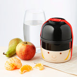 In-context lifestyle shot of a Bento Round Stacked Lunch Box, front view, styled as a London Guardsman with a black helmet-like top and a beige face base, secured by a red strap. It sits on a woven placemat next to an apple, a pear, a glass of water, and peeled satsuma segments.