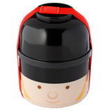 Front view of a round, stacked bento lunch box styled as a London Guardsman. It features a shiny black top, a beige lower section with a stylised face, brown eyes, a smile, and a yellow uniform stripe. A red fabric strap secures the two tiers.
