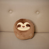 Front view of a brown plush squeezies sloth cushion, embroidered with a face showing eyes, heart-shaped nose, mouth, and claw markings, on a beige upholstered sofa.