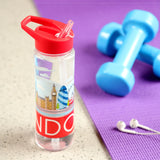 Angled view of a 550ml reusable water bottle featuring a colourful London skyline illustration and red 'LONDON' text. The bottle has a red flip straw lid and is pictured on a grey surface next to a purple mat, blue dumbbells, and white earphones.
