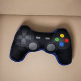 Overhead view of a black plush game controller cushion with blue trim, featuring a grey D-pad, grey joysticks, and colourful red, blue, yellow, and green buttons, resting on a beige surface.