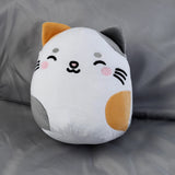 Front view of a white plush cat toy with grey and orange patches, grey and orange ears, and an embroidered smiling face with pink cheeks, on a grey fabric background.