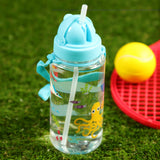 Angled view of a clear children's water bottle with a light blue straw lid and strap, decorated with colourful sealife creatures including a yellow octopus, on green grass with a red tennis racket and yellow ball in the background.
