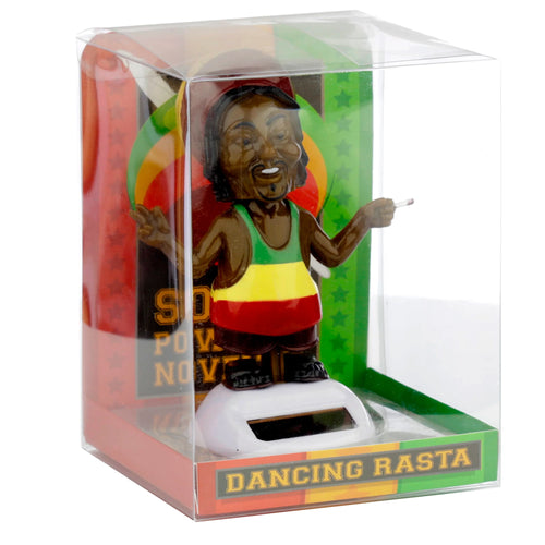 Angled view of a Rasta Solar Pal figurine in clear retail packaging. The smiling figure has dreadlocks, a beard, and wears a green, yellow, and red vest. It gestures a peace sign with one hand and holds a white object in the other, standing on a white base with a solar panel. The packaging displays a colourful backing card and 'DANCING RASTA' text on the front.
