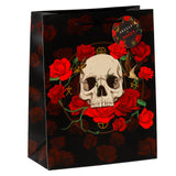 Angled front view of a large black gift bag featuring a prominent design of a white skull encircled by vibrant red roses, green thorny vines, a moth, a flying bird, and golden symbols. It has black rope handles and a circular 'Skulls + Roses' tag.