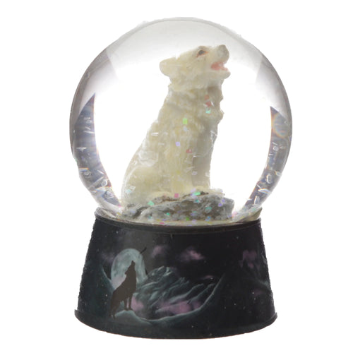Front view of a snow globe featuring a white wolf figurine howling, surrounded by iridescent glitter. Its dark base depicts a silhouetted wolf howling at the moon against mountains under a night sky.