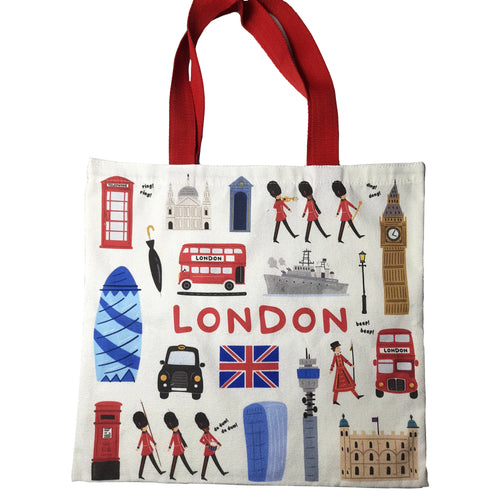 Front view of an off-white canvas London souvenir shopping bag with red handles, featuring a colourful cartoon design of iconic London landmarks including Big Ben, red double-decker buses, telephone boxes, Beefeaters, and the Union Jack flag.
