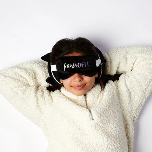 In-context lifestyle shot, overhead view of a person relaxing in a cream fluffy jumper, wearing a black plush cat ear travel pillow and a black 'pawsOFF!' eye mask.