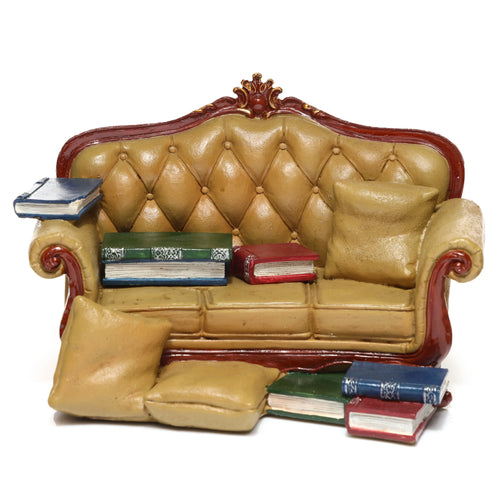 Angled view of a miniature tan sofa-shaped display stand with a tufted backrest, ornate reddish-brown and gold trim, featuring small decorative blue, green, and red books with matching cushions.