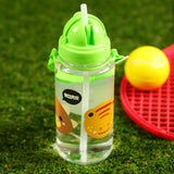 In-context lifestyle shot of a clear water bottle featuring colourful dinosaur illustrations, including a brown character, a yellow spiky character, and a 'ROAR' speech bubble. It has a bright green flip-top lid, an integrated white straw, and a green carry strap, set on green grass with a blurred red toy racket and yellow ball in the background.