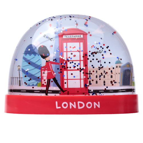 Front view of a London-themed snow globe. The red base displays 'LONDON' in white. Inside the clear dome, a British Guardsman in red uniform marches past a red telephone box, with the Gherkin and other London buildings in the background, amidst red, white, and blue glitter.