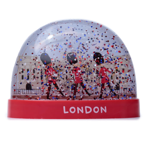 Front view of a snow globe with a red base displaying 'LONDON' in white text. Inside, three marching British guards in red uniforms and black bearskin hats are positioned in front of a light-coloured building, surrounded by red, blue, and white glitter.
