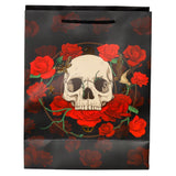 Front view of a large black gift bag with black rope handles, featuring a central graphic of a light-colored skull surrounded by vibrant red roses and green vines. Additional darker red rose patterns are visible in the background, along with subtle gold accents including a moth and a flying bird.