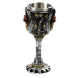 Angled view of a decorative steampunk skull goblet with a distressed grey and silver metallic finish and brass accents. The goblet features skull heads wearing top hats, gears, and pipe details on the bowl, a screw-thread stem, and a cog-shaped base.