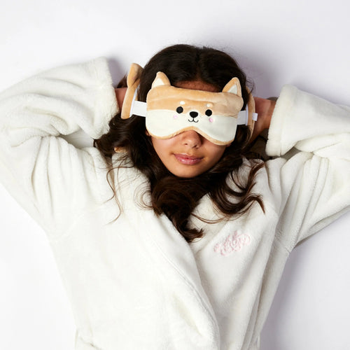 In-context lifestyle shot from above of a person wearing a beige plush Shiba Inu dog eye mask with embroidered features and small ears, with the matching plush travel pillow behind their head, lying down in a fluffy white robe.