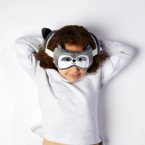 Overhead lifestyle shot of a person wearing a grey raccoon eye mask with small ears, lying relaxed and using a matching plush raccoon travel pillow on a white background.