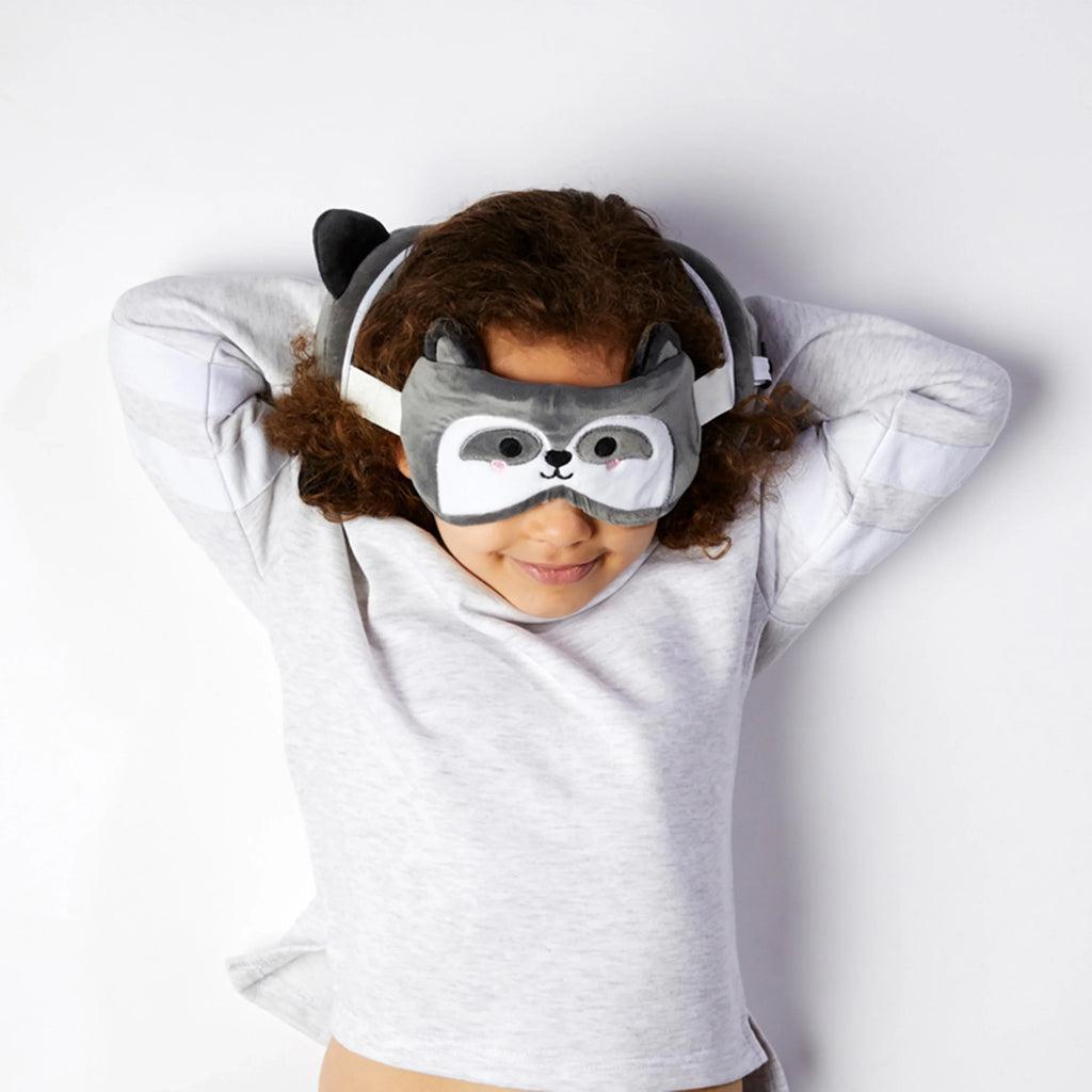 Overhead lifestyle shot of a person wearing a grey raccoon eye mask with small ears, lying relaxed and using a matching plush raccoon travel pillow on a white background.