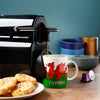 In-context lifestyle shot featuring a white porcelain mug decorated with a red Welsh dragon and 'CYMRU' text on a green band, receiving steaming coffee from a machine. A plate of biscuits and a purple coffee pod are visible on the wooden counter.