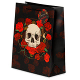 Angled front view of a large black gift bag, featuring a prominent graphic of a white skull encircled by bright red roses. The bag is also patterned with darker red roses, and it has black cord handles with a circular 'Skulls + Roses' tag attached.