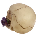 Left profile of a light beige Gothic skull decoration with prominent dark cracks, holding a vibrant purple rose with green leaves in its jaw.