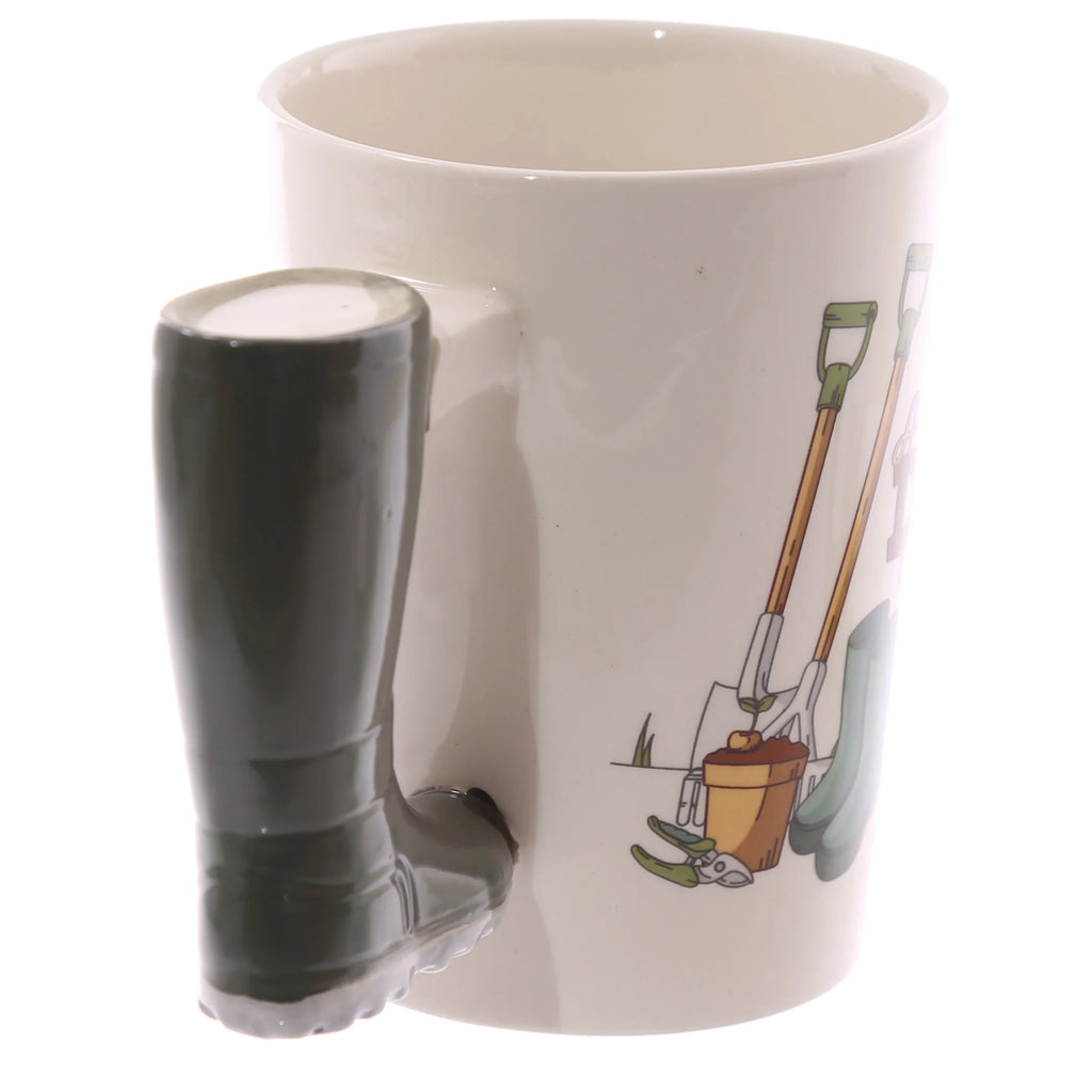 Angled view of a cream ceramic mug with a dark green Wellington boot handle and colourful garden tool illustrations.