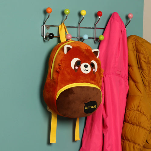 Front view of an orange and brown red panda plush rucksack backpack with yellow straps and zipper, hanging on a colorful coat rack mounted on a teal wall, alongside a pink jacket and a mustard jacket.