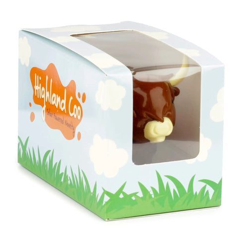 Angled view of a Collectable Highland Coo Cow Solar Powered Pal in its packaging. The brown cow figure, with white horns and a light snout, is visible through the clear window of the light blue box, decorated with grass and clouds.