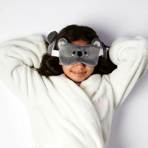Overhead lifestyle shot of a smiling person wearing a grey plush koala eye mask with embroidered features and ears. The matching koala travel pillow is visible behind their head, on a white background.