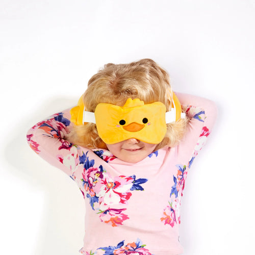 Lifestyle shot of a child with blonde hair wearing a plush yellow duck eye mask with black eyes and an orange beak, part of a travel pillow set. The child wears a pink floral top.