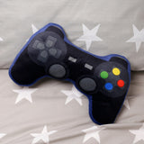 Overhead view of a black plush game controller cushion with blue trim, featuring colourful buttons and grey controls, lying on a grey fabric with white star patterns.