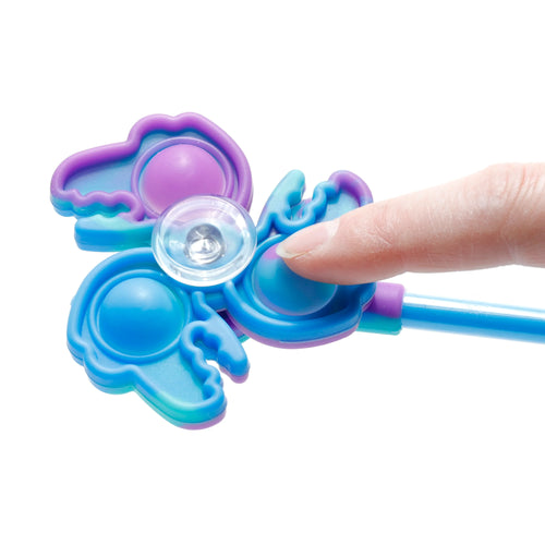Angled close-up of a multicoloured (blue, turquoise, purple) kids' spinner fidget top pen. A finger presses a pop-it bubble on the dinosaur and unicorn-themed fidget toy, demonstrating its use.