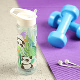 Lifestyle shot of a clear reusable water bottle with a white flip-straw lid, featuring a panda and green foliage design. The bottle is angled on a light surface, with blurred blue dumbbells and white earphones on a purple yoga mat in the background.
