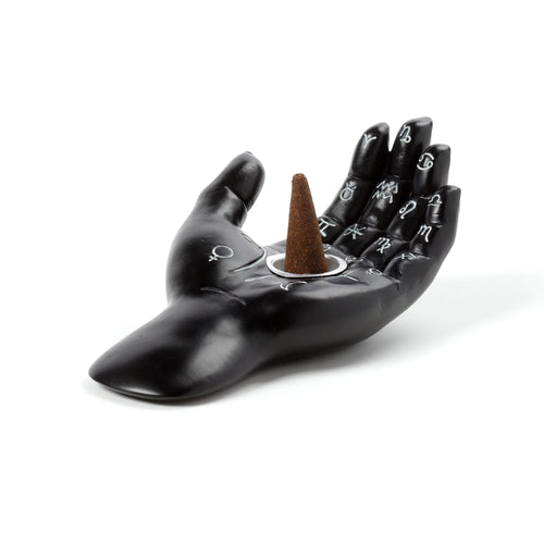 Angled view of a black decorative hand-shaped incense cone dish, featuring white astrological and zodiac symbols etched across the palm and fingers, holding a brown incense cone in its silver holder.