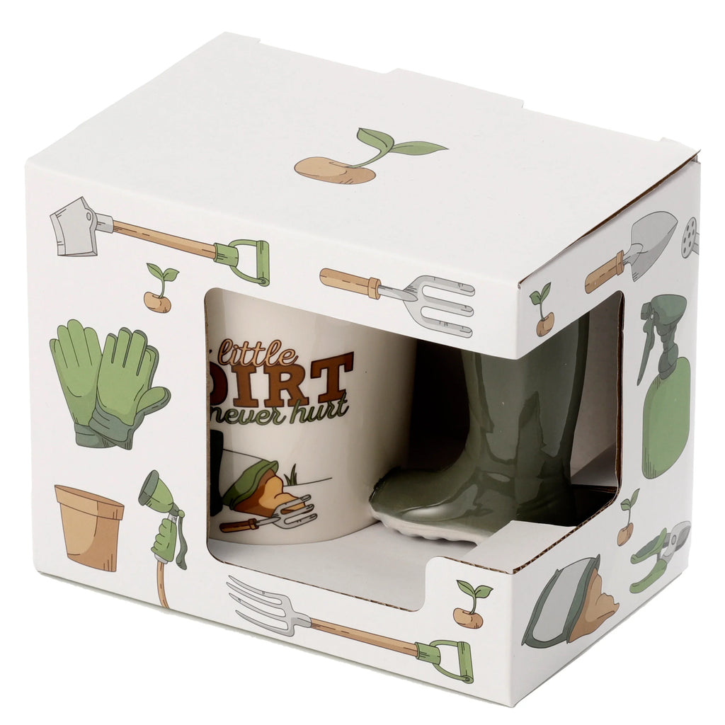Angled view of a cream ceramic mug featuring 'little DIRT never hurt' text and a gardening illustration, with its olive green Wellington boot handle visible through a cut-out in white cardboard packaging decorated with various gardening tool illustrations.
