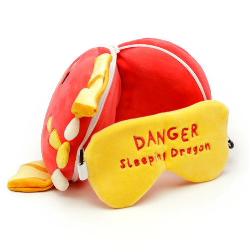 Angled view of a red plush dragon travel pillow with yellow wing details, next to a yellow eye mask embroidered with 'DANGER Sleeping Dragon'.