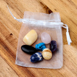 Overhead view of a Chakra Stones Mini Set of 7, featuring seven polished stones in various colors visible inside a white sheer drawstring pouch, resting on a wooden surface.