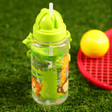 In-context lifestyle shot: Clear children's water bottle with a bright lime green lid, exposed straw, and green fabric carry loop. The bottle displays a colourful zoo animals design featuring a green crocodile, yellow giraffe, brown monkey, and orange tiger. It rests on green artificial grass, with a blurred red tennis racket and yellow ball in the background.