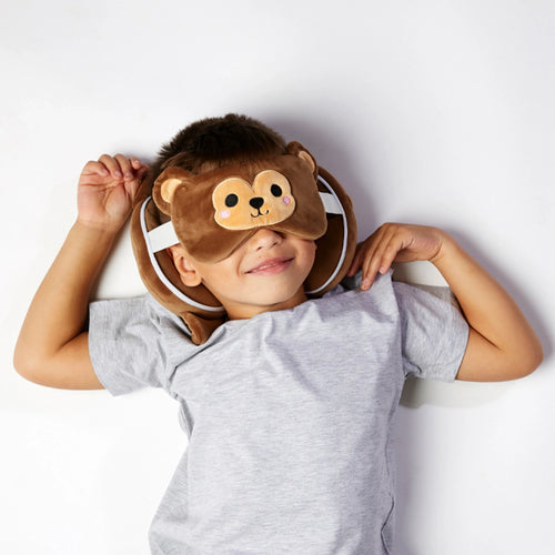 Overhead lifestyle shot of a child wearing a brown monkey plush travel pillow and matching monkey face eye mask, smiling whilst lying down.