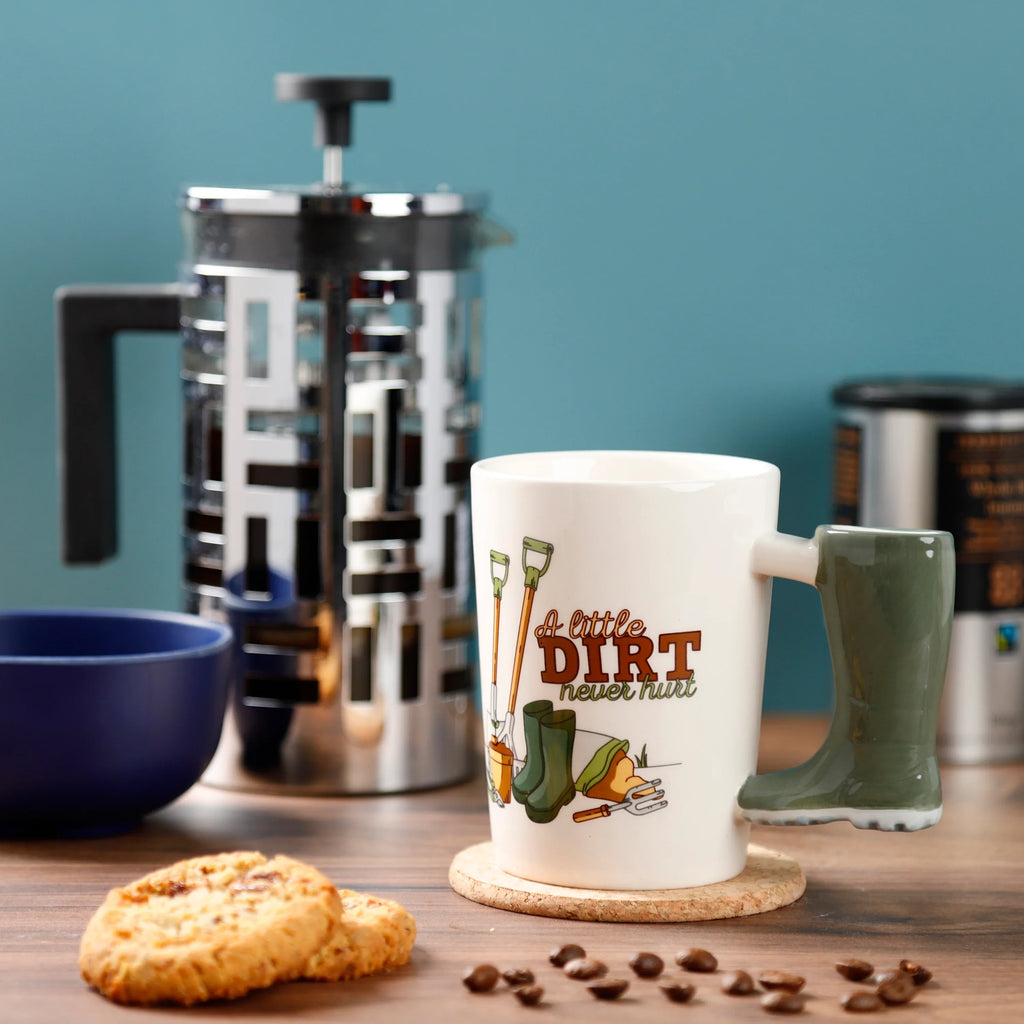 Angled view of a cream ceramic mug with a green Wellington boot-shaped handle. The mug body features 'A little DIRT never hurt' text and gardening illustrations. It is placed on a cork coaster, surrounded by scattered coffee beans and two cookies, with a chrome cafetiere visible in the background.