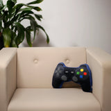 Blue and black plush game controller cushion, showing its D-pad and colourful buttons, resting on a cream armchair.