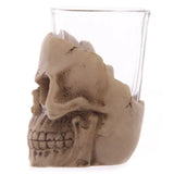 Left-side profile view of a beige skull mini shot glass, featuring a clear glass insert and an aged appearance with dark accents on the eye socket, nose cavity, and teeth.